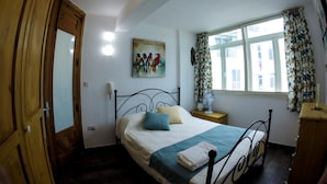 Ein Schlafzimmer mit einem Bett, einem Nachttisch, Vorhängen an einem Fenster, einem Bild an der Wand und einem Holzschrank.
