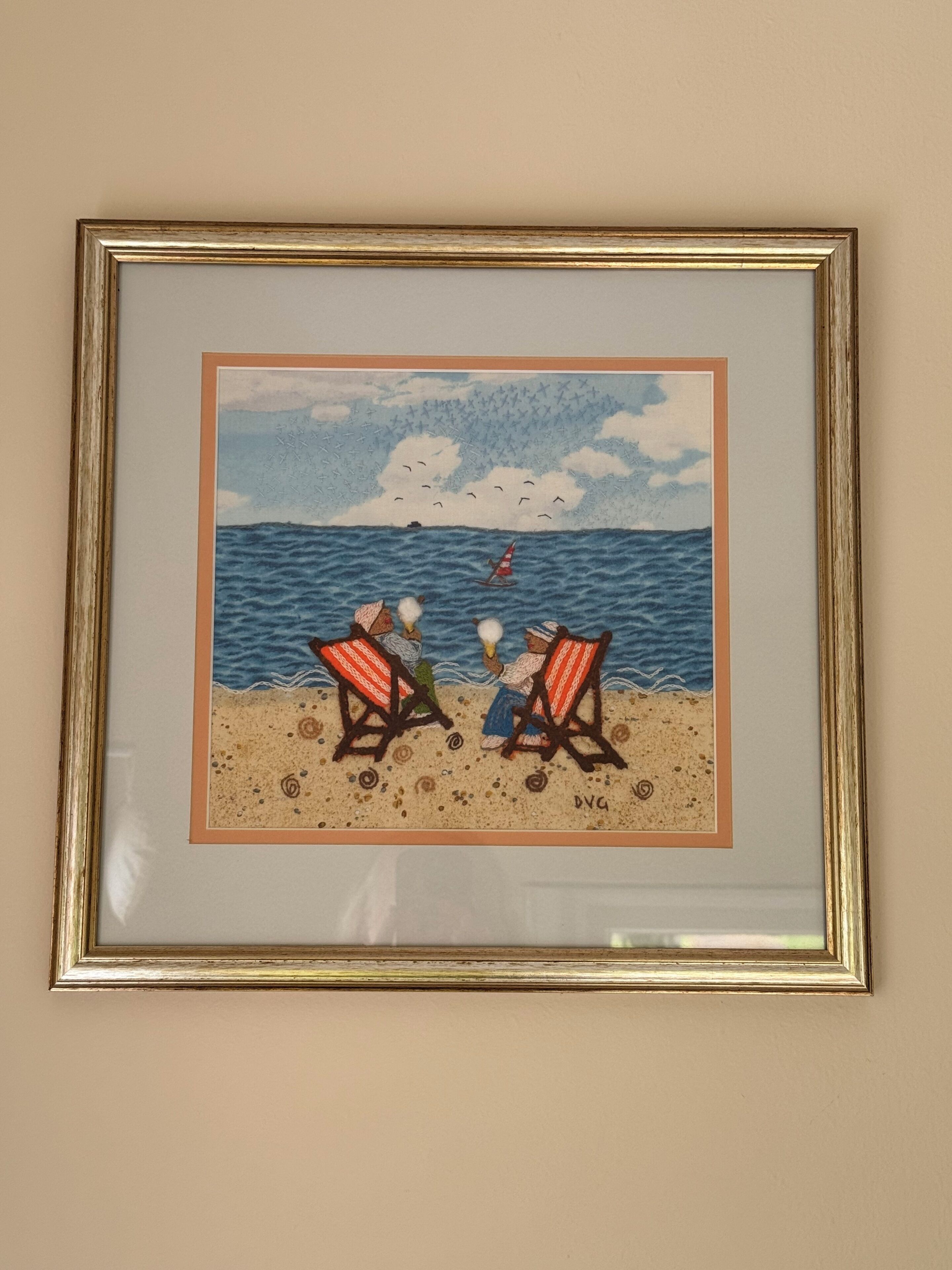 A framed artwork depicting two figures on a beach with a sailboat and birds in the sky.