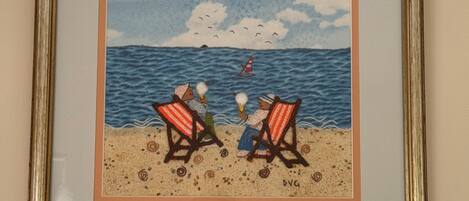 A framed artwork depicting two figures on a beach with a sailboat and birds in the sky.