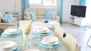 A dining area with a glass table set for a meal, a sofa, a TV, and a window with curtains.