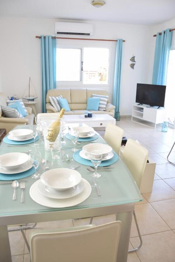 Un coin repas avec une table en verre mise pour le repas, un canapé, un téléviseur et une fenêtre avec des rideaux.