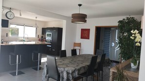 A modern kitchen with dark cabinetry, a built-in oven, and a large island with bar stools. There is a dining area with a patterned tablecloth and chairs. - superb house ideally located 220 m2 (Sainte-Sève)