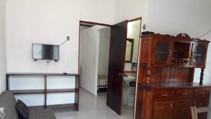 Una sala de estar con un sofá, un televisor montado en la pared, un mueble de madera y una puerta que da a otra habitación.
