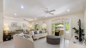 Un salon spacieux avec un canapé beige, un pouf rond et un ventilateur de plafond.