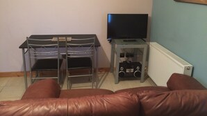 A living room with a brown leather sofa, a TV on a stand, a metal shelf unit, and a clock on the wall. - 1 Bedroom Chalet 30 Minute Walk From Stornoway Town Centre (Stornoway Isle of Lewis)
