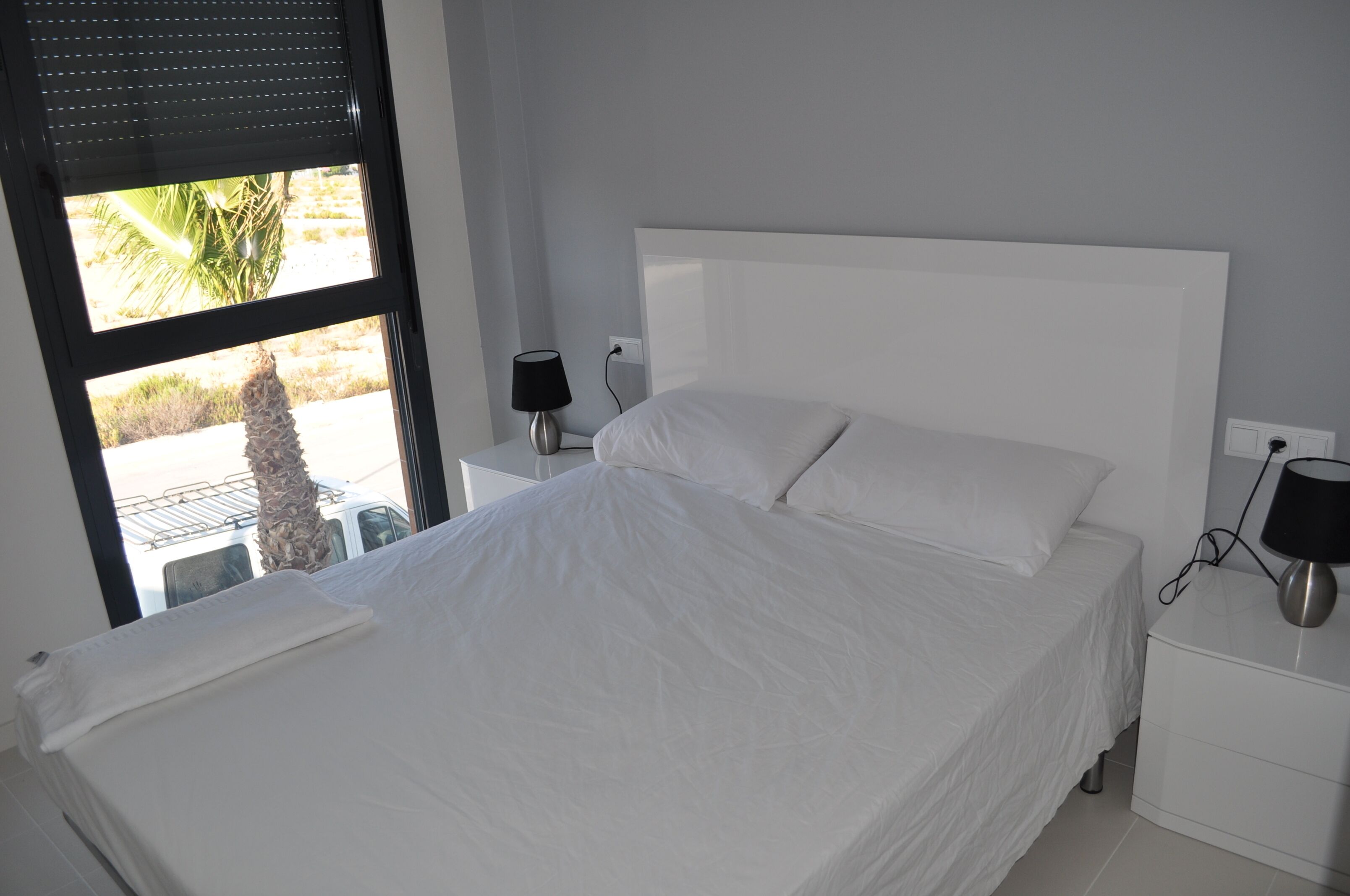 A neatly made bed with white linens, a white nightstand with a lamp, and a window with blinds.