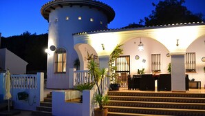 A white building with a round tower, a staircase leading to a covered patio area, and outdoor seating.