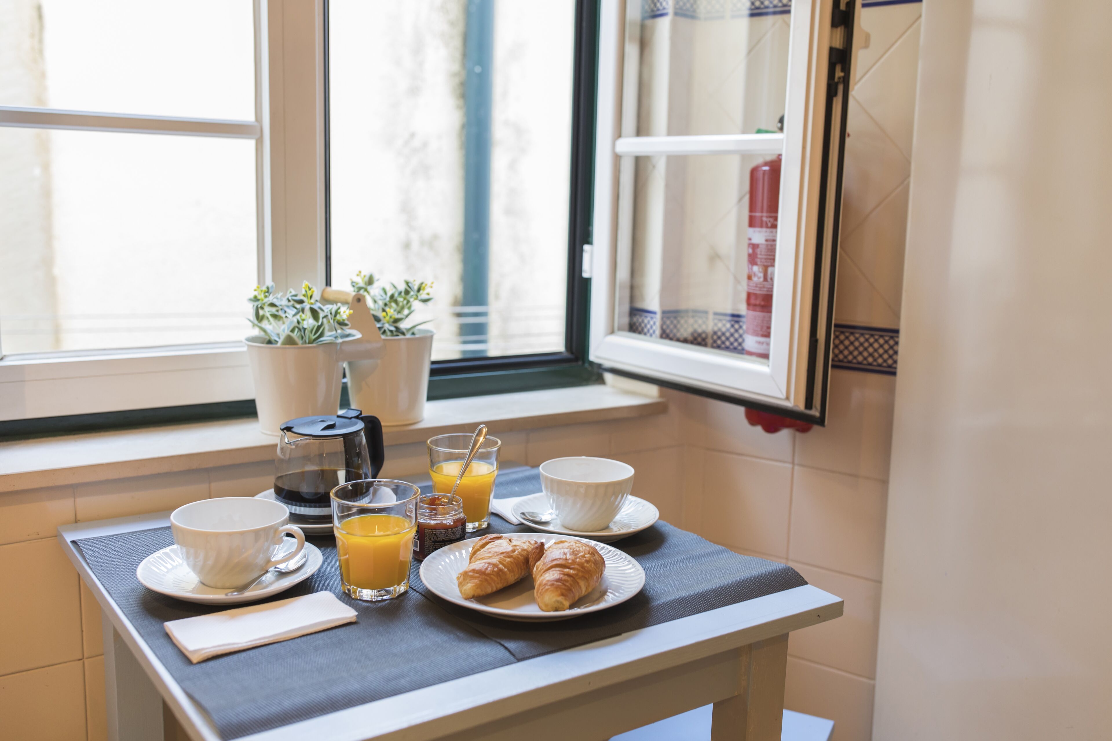 A breakfast table with croissants, orange juice, and coffee, set by a window with potted plants.