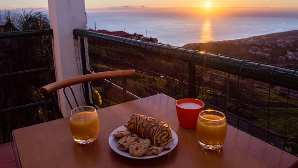 A table with a plate of cookies and two glasses of orange juice, a candle, and a view of the sunset over the ocean. - Wonderful View Apartment with Fantastic sunsets (Florenças, Arco da Calheta)