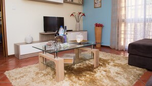 A modern living room with a glass coffee table, a flat-screen TV, a white cabinet, and a beige rug. - Wonderful View Apartment with Fantastic sunsets (Florenças, Arco da Calheta)
