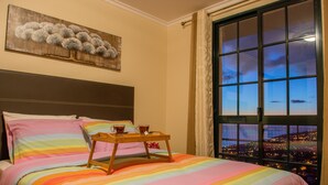 A bed with a rainbow-striped bedspread, a wooden tray with two cups, and a cityscape view through a window. - Wonderful View Apartment with Fantastic sunsets (Florenças, Arco da Calheta)