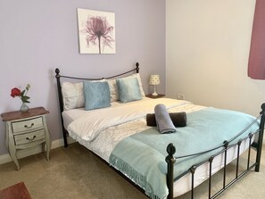 A neatly made bed with a blue and white bedspread, a decorative pillow, and a rolled towel. There is a small bedside table with a vase of flowers and a lamp.