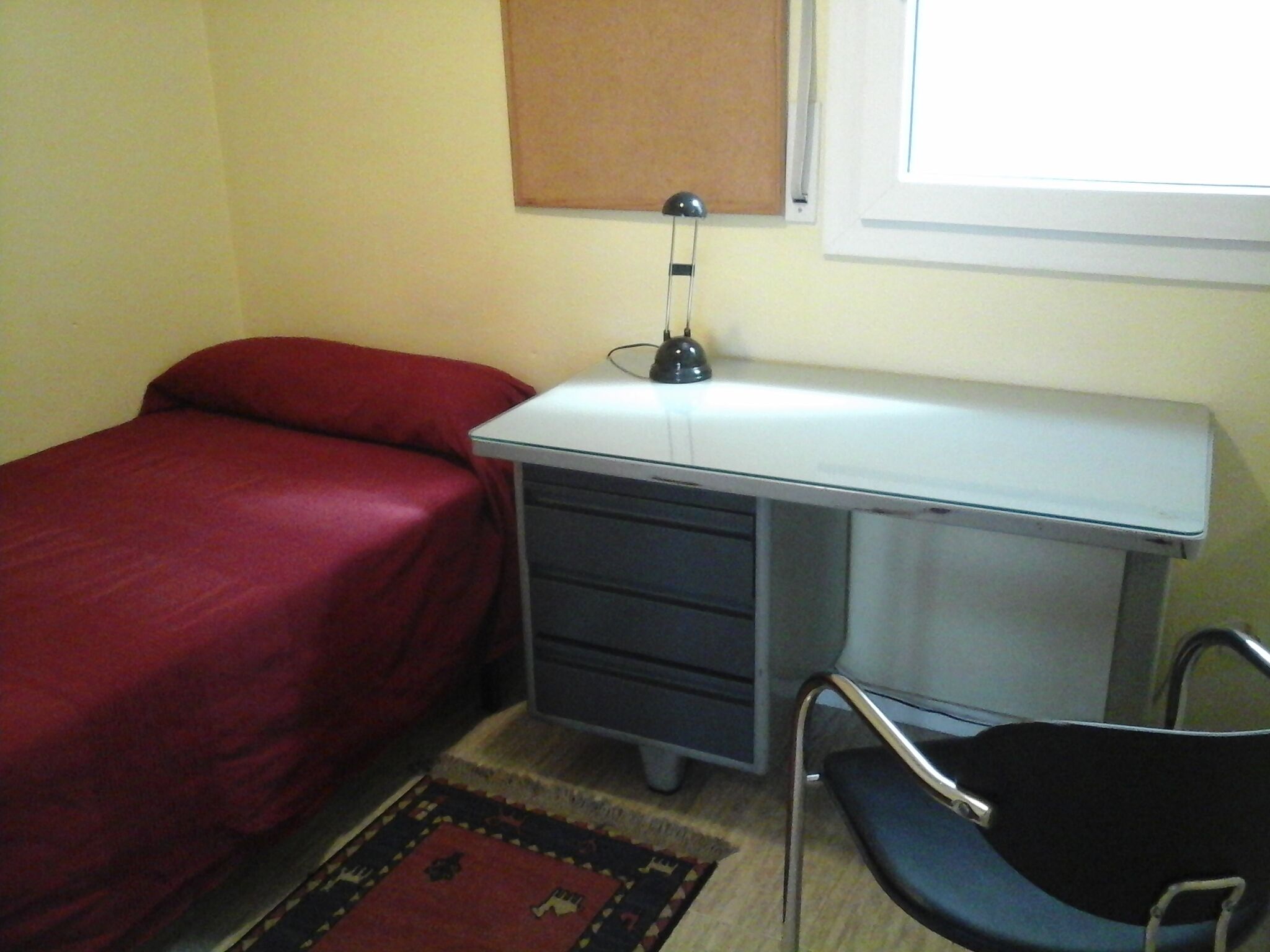 Een kleine slaapkamer met een enkel bed, een bureau met lades, een stoel, een lamp en een raam.