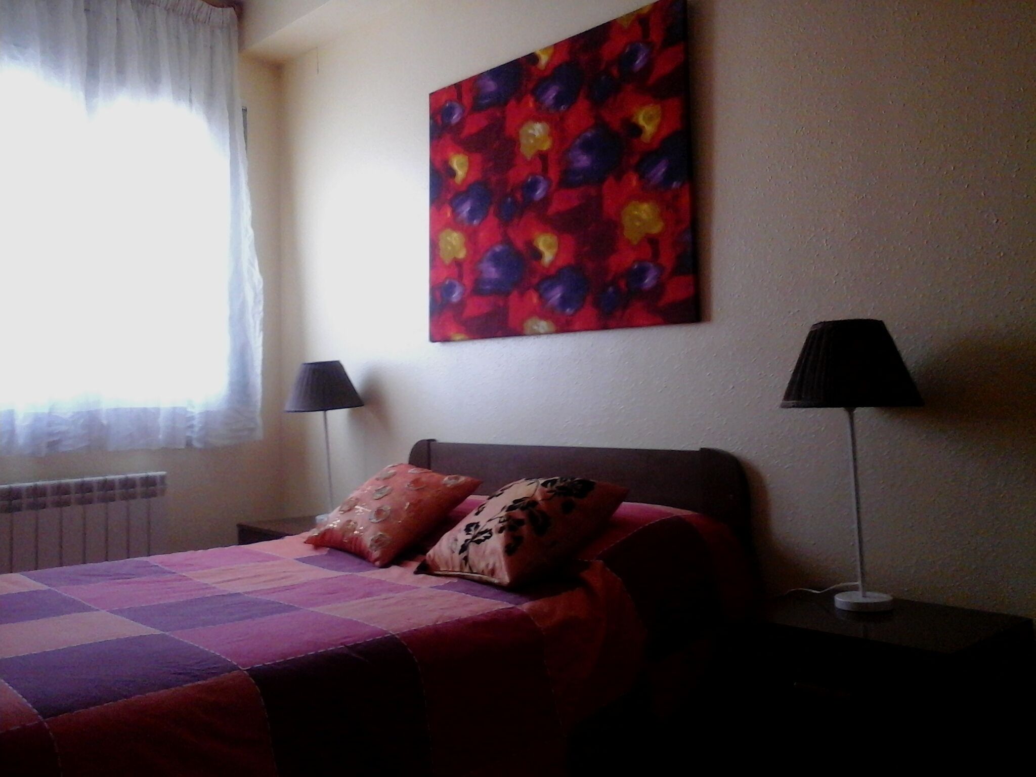 Quarto com cama, duas luminárias de cabeceira, radiador e um quadro abstrato colorido na parede.