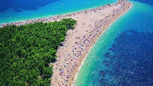 Ovenudblik over en strand med en tæt skov på den ene side og en sandstrand med mange parasoller og mennesker.
