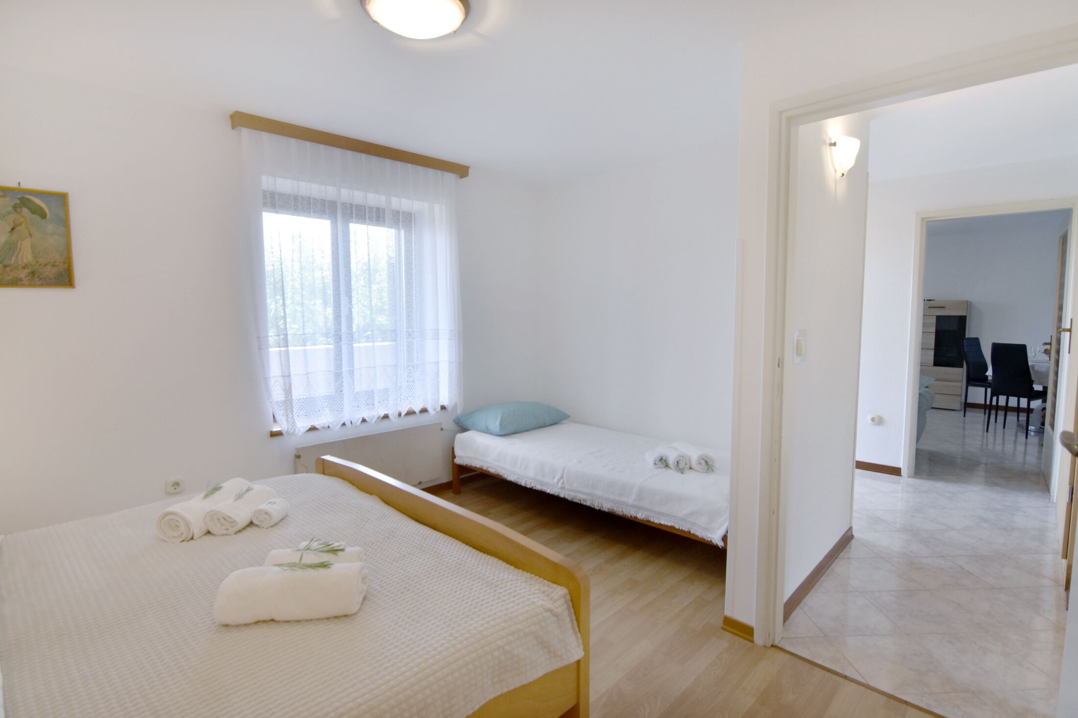 A room with two beds, one with a wooden frame and the other with a metal frame. There is a window with sheer curtains, a painting on the wall, and a door leading to another room.