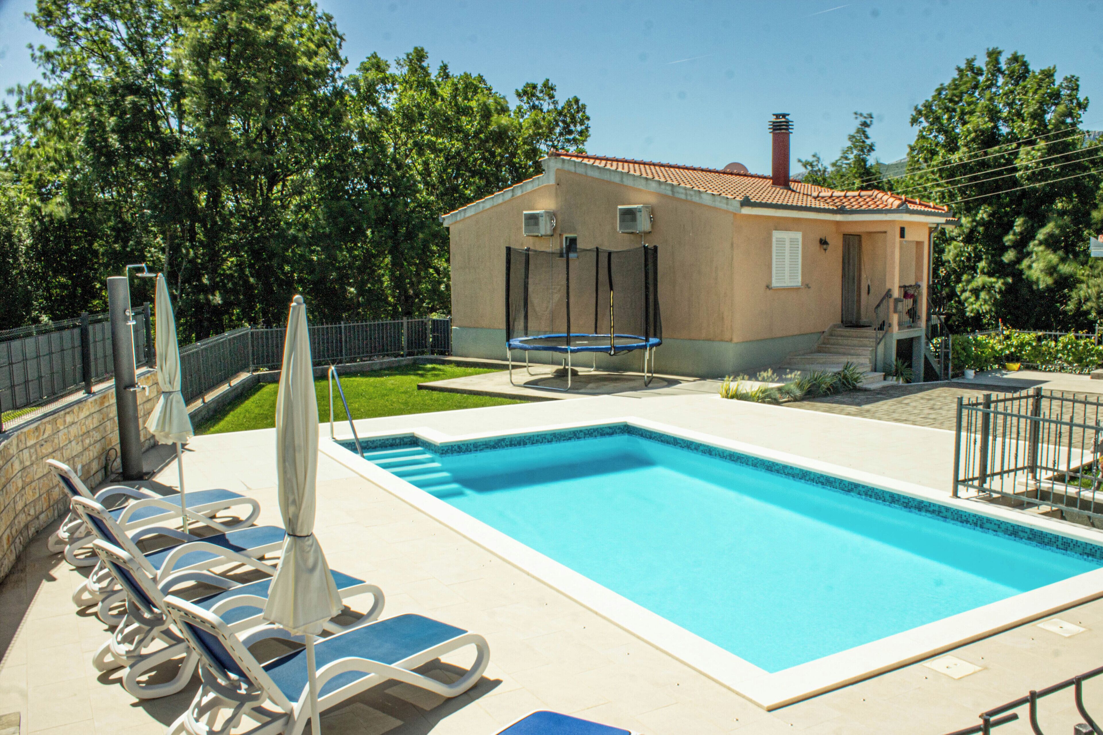 A swimming pool with lounge chairs and umbrellas, a small building with a red roof, and a fenced area with trees.