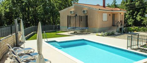 A swimming pool with lounge chairs and umbrellas, a small building with a red roof, and a fenced area with trees.