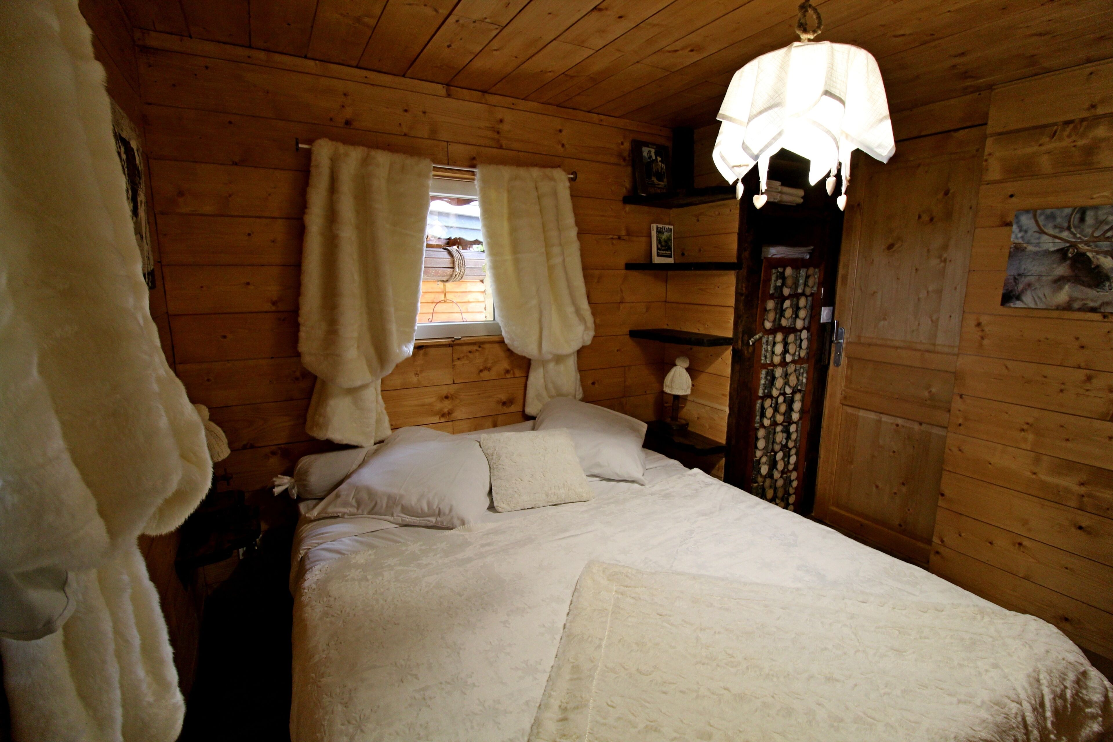 Camera da letto in una baita di legno, con un letto, tende, un comodino, una lampada e una porta con pannello decorativo.