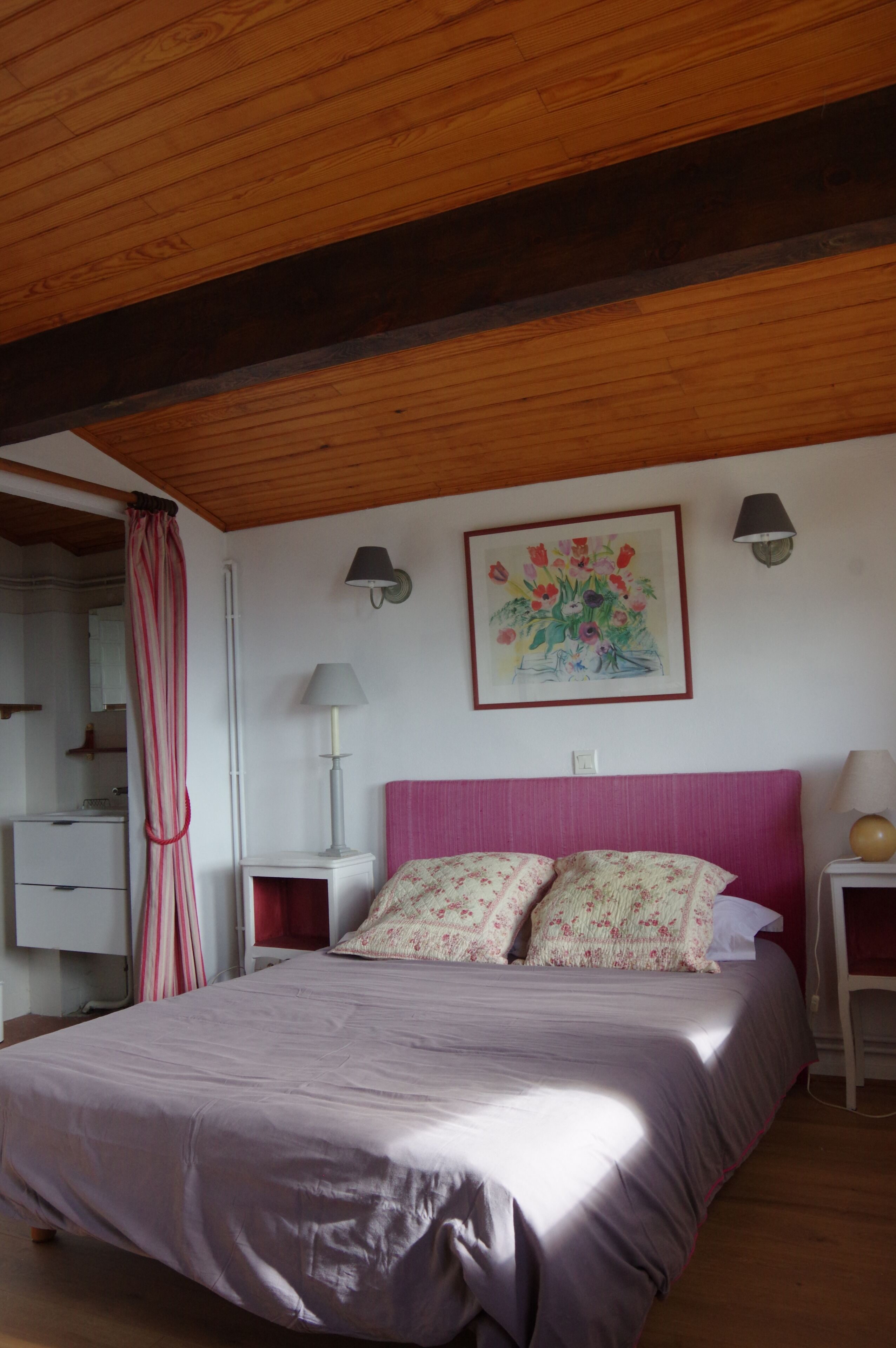 Une chambre à coucher comprenant un lit, une table de chevet, une lampe, un luminaire fixé au mur, un tableau et une fenêtre avec des rideaux.