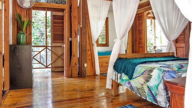 Une chambre dans une cabane en bois, avec un lit, des rideaux, un ventilateur et une vue sur l’extérieur.