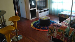 Una stanza con pavimento in legno, uno sgabello giallo, un tavolo rotondo, una TV su un mobile e un letto con copriletto colorato.