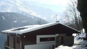 A chalet with a stone foundation, wooden exterior, and a snow-covered roof. It is situated on a snow-covered slope with mountains in the background.