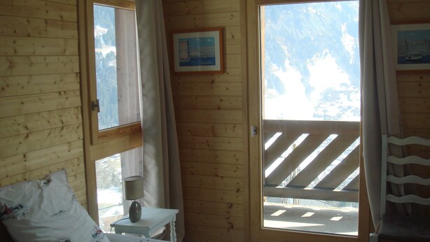 A wooden cabin room with a bed, a nightstand, a window with curtains, and a view of a snowy landscape.
