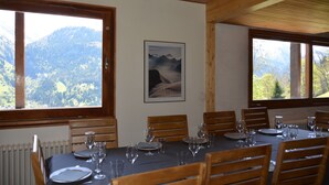 A dining area with a table set for six, large windows with mountain views, wooden ceiling beams, and a framed picture on the wall.