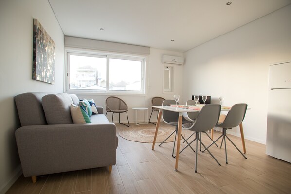 Un salon moderne avec un canapé, une table à manger et des chaises. Une fenêtre laisse entrer la lumière naturelle et offre une vue sur les bâtiments environnants.