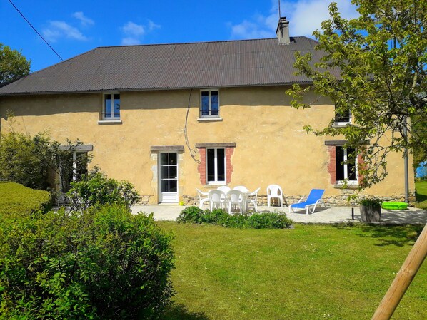 Une maison à deux étages avec un toit plat, une véranda à l’avant avec une table et des chaises, et un petit jardin.