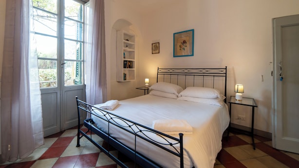 A bedroom with a bed, a window with curtains, a bedside table with a lamp, and a door. - Studio in the heart of Finalborgo (Finale Ligure)
