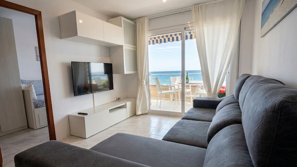 A modern living room with a large flat-screen TV, grey sofas, a sliding glass door leading to a balcony with a view of the sea. - Spectacular Frontline, Pineda de Mar (Pineda de Mar)