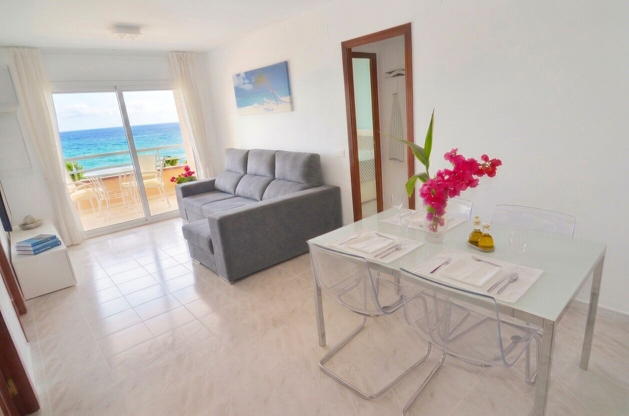 A room with a balcony, a grey sofa, a glass dining table with chairs, and a vase with flowers.
