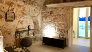 Una habitación rústica con paredes de piedra, una cama en altillo, una mesita con una planta y un banco.