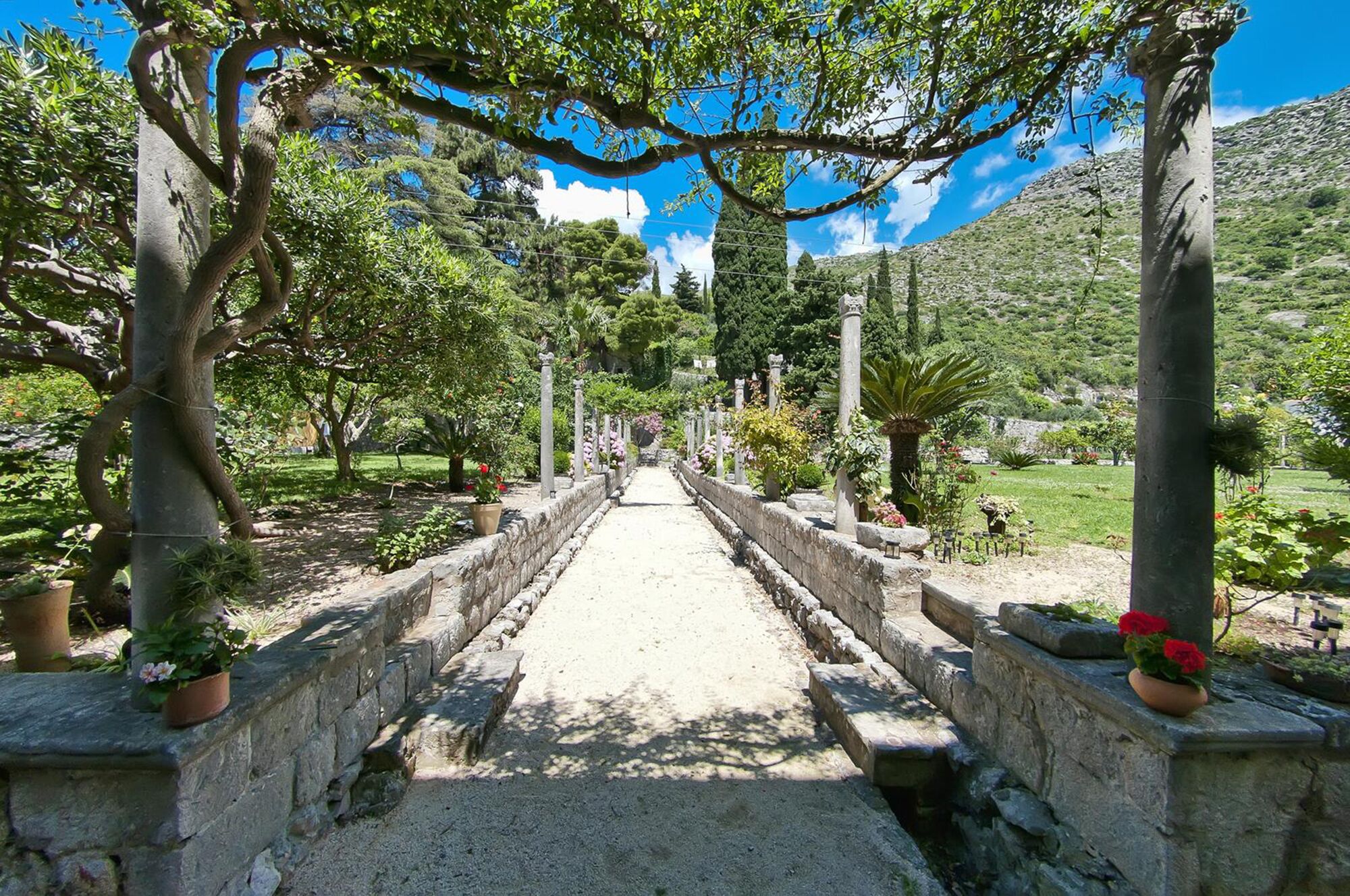 A pathway flanked by stone walls and potted plants, leading through a garden with trees and a mountainous backdrop.