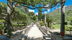 Un sendero bordeado por muros de piedra y macetas con plantas, que atraviesa un jardín con árboles y montañas al fondo.