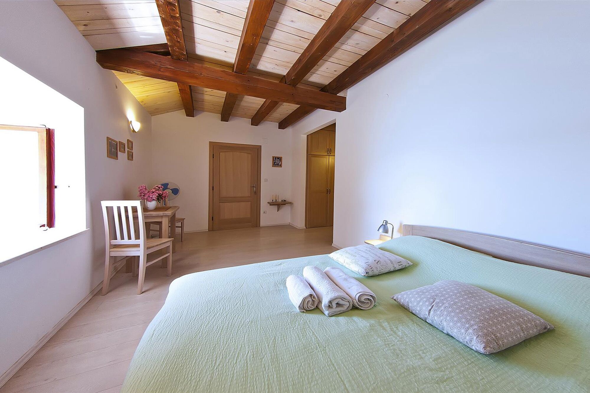 A bedroom with a bed, a chair, a table with flowers, and a door leading to another room.