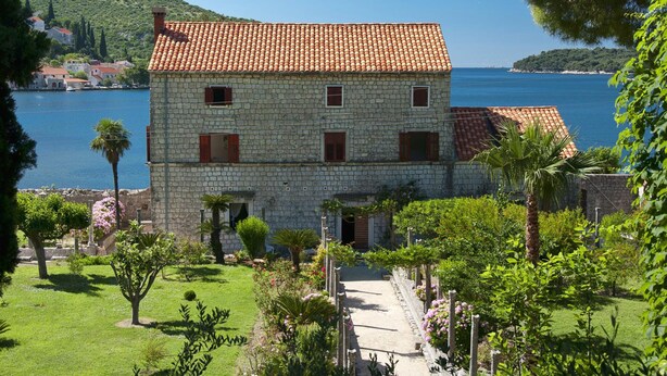 Un edificio de piedra con techo de tejas rojas, un jardín con diversas plantas y vistas al mar.