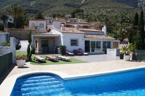 En vy över en villa i medelhavsstil med en pool, solstolar och en uteplats, allt sett från poolområdet.