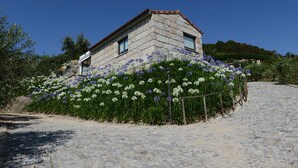 Een stenen huis met een pannendak, omringd door een tuin vol witte en paarse bloemen, en een keienpad dat er naartoe leidt.