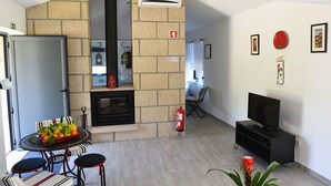 Een woonkamer met een stenen openhaard, een eethoek met een tafel en stoelen, een televisie op een meubel en een brandblusser op de vloer.