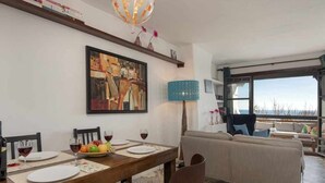 Un coin repas avec une table en bois, des chaises et un panier de fruits. Il y a un tableau au mur et un canapé à l’arrière-plan.