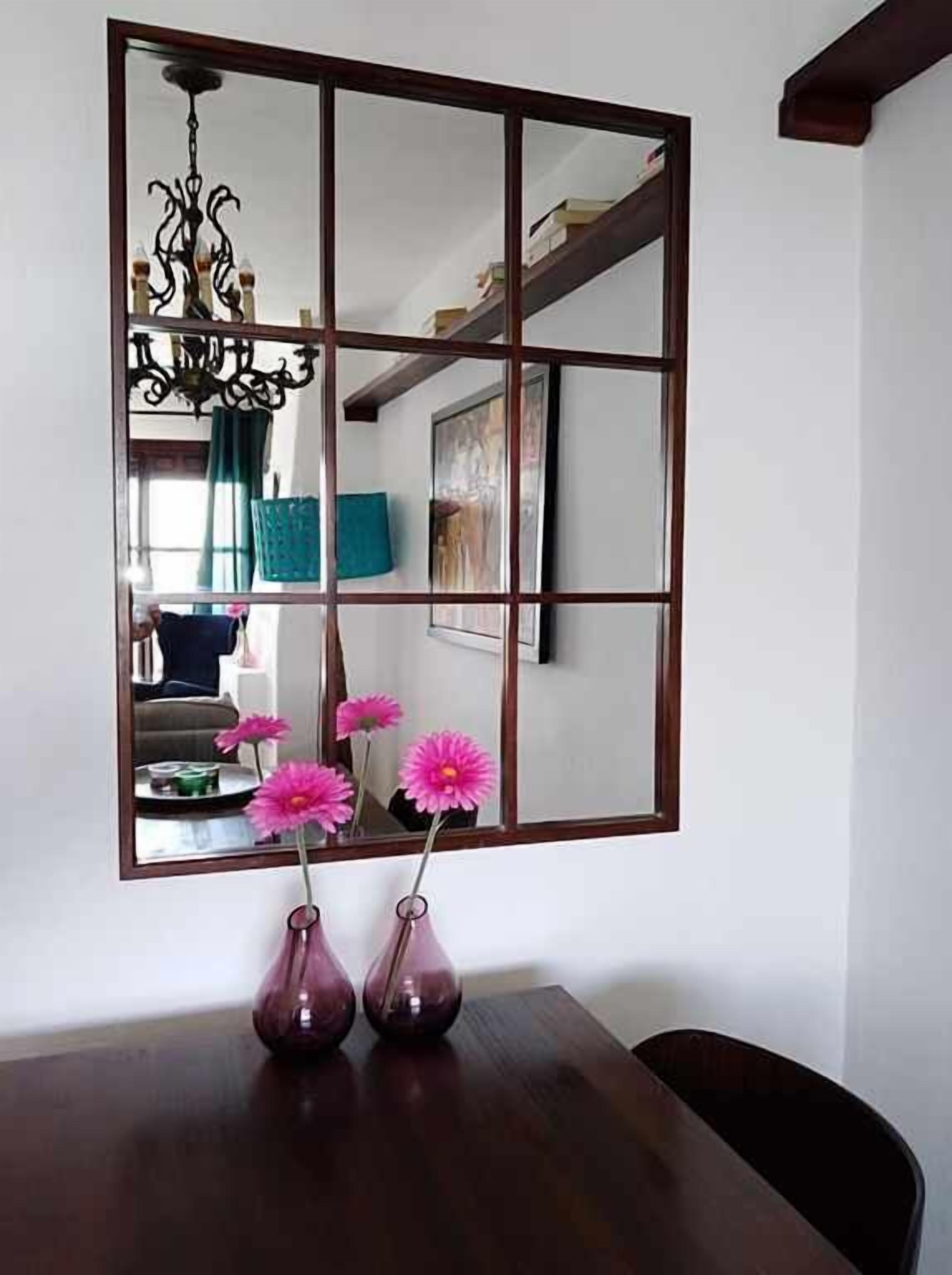 Un comedor con una mesa de madera, tres jarrones con flores rosas y un espejo grande que refleja la habitación.