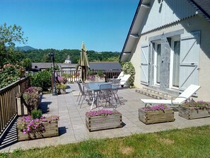 A patio with a table and chairs, surrounded by wooden planters with flowers, and a building with large windows and a door. - Bed and breakfast at the farm, Lourdes, Pyrenees, ANOUSTA (Loubajac)
