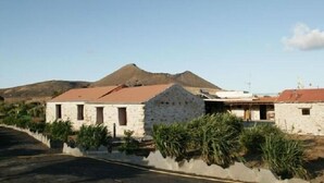 A stone building with a red roof, surrounded by greenery and a mountain in the background.
