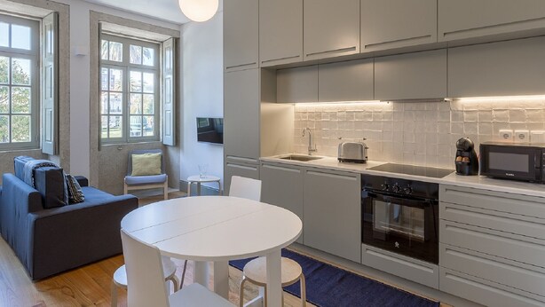 Eine moderne Küche mit grauen Schränken, einem Backofen, einer Mikrowelle und einer Kaffeemaschine. Es gibt einen runden Esstisch mit Stühlen, eine Couch und einen an der Wand befestigten Fernseher.