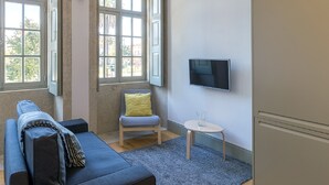 Ein modernes Wohnzimmer mit einer Couch, einem kleinen Tisch, einem an der Wand befestigten Fernseher und großen Fenstern mit Blick nach draußen.