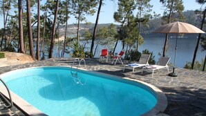 A swimming pool with a ladder, surrounded by stone tiles, and overlooking a lake with chairs and an umbrella.
