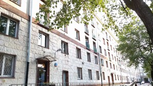 Un edificio de varios pisos con fachada de piedra, una entrada visible con toldo metálico y una calle con automóviles estacionados.